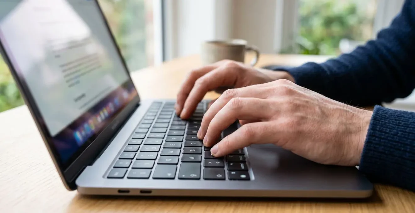 Nahaufnahme von Händen, die auf einer Laptop-Tastatur tippen, unscharfer Bildschirm mit Daten im Hintergrund, helles Tageslicht